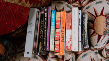 A stack of 14 personal growth and self-help books featured in a summer 2025 reading list, laid on a sun-patterned woven blanket.