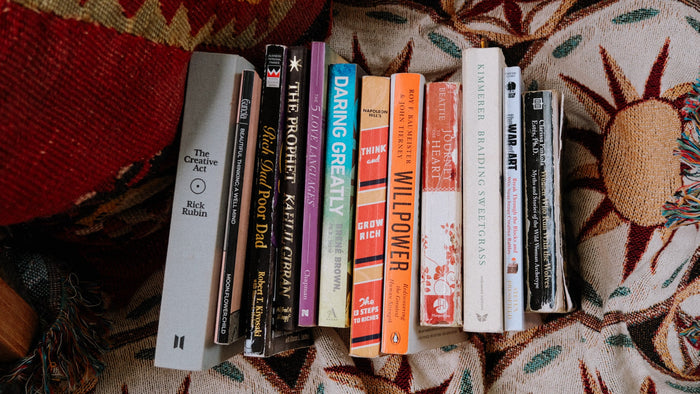 A stack of 14 personal growth and self-help books featured in a summer 2025 reading list, laid on a sun-patterned woven blanket.