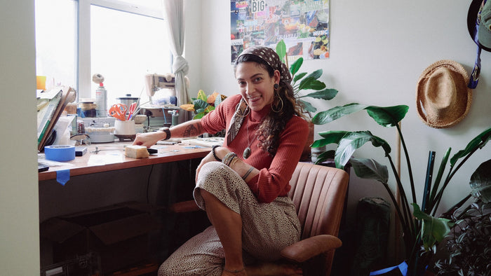Woman sitting at a cozy workspace, smiling warmly at the camera. She is dressed in a rust-colored sweater, patterned pants, and a headscarf, with statement jewelry. The desk is surrounded by plants, and the room has a creative, inviting atmosphere